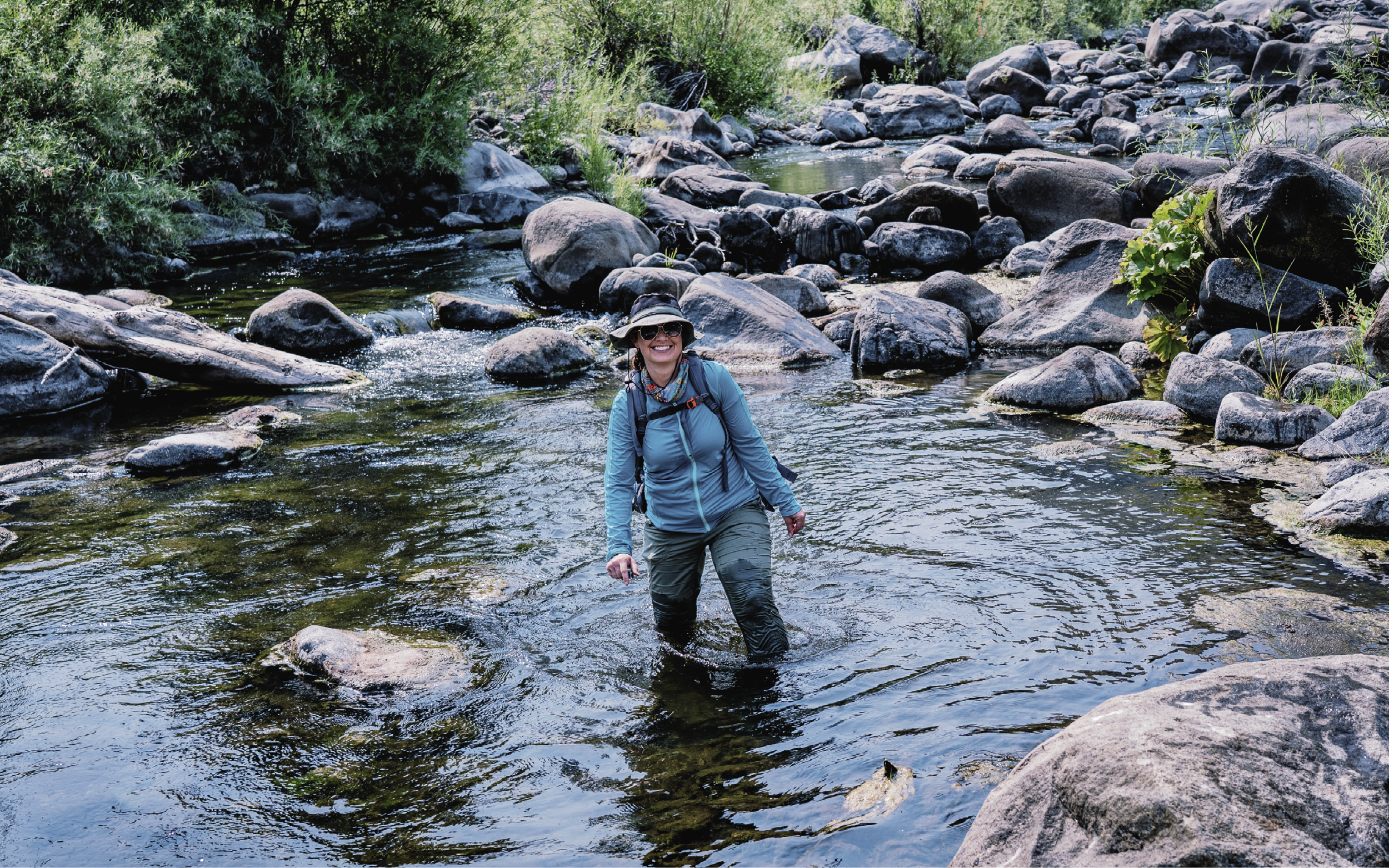 Claudia Mengelt is wearing blue waders and is knee-deep in a rocky river, smiling at the camera.
