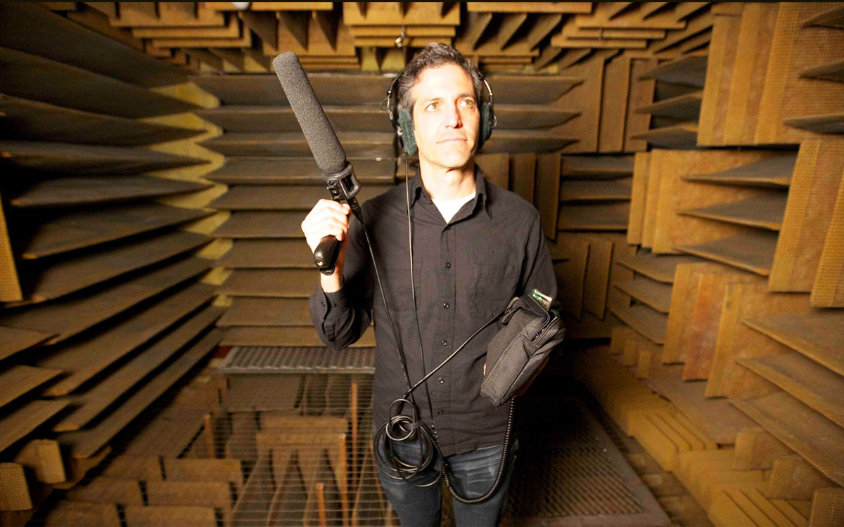 A man in headphones holding a shotgun mic and reorderstands in a low-ceilinged room covered in blocks of orange soundproofing foam.