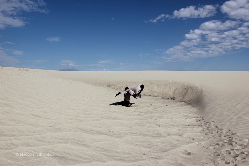 A self portrait of Blanca taken on a sand dune on a sunny day. There are clouds on the upper left and right edges of the image and a slight view of a mountain on the horizon. Blanca, a brown femme, sits in the center, staring directly at the viewer.