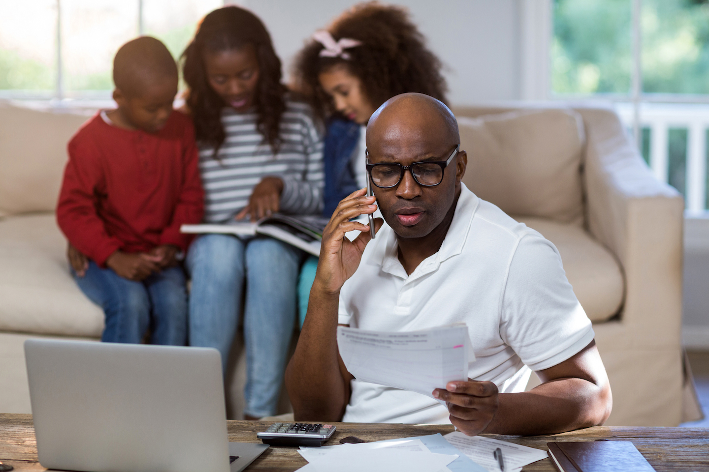 Man talking on a mobile phone in front of a laptop and papers, with his family sitting on a couch behind him