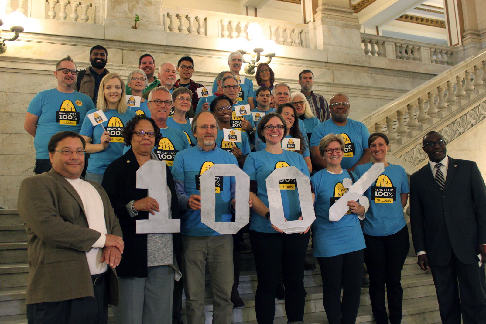 A group of people wearing blue Ready for 100% shirts stand on marble steps and hold up lettering that spells 100%.