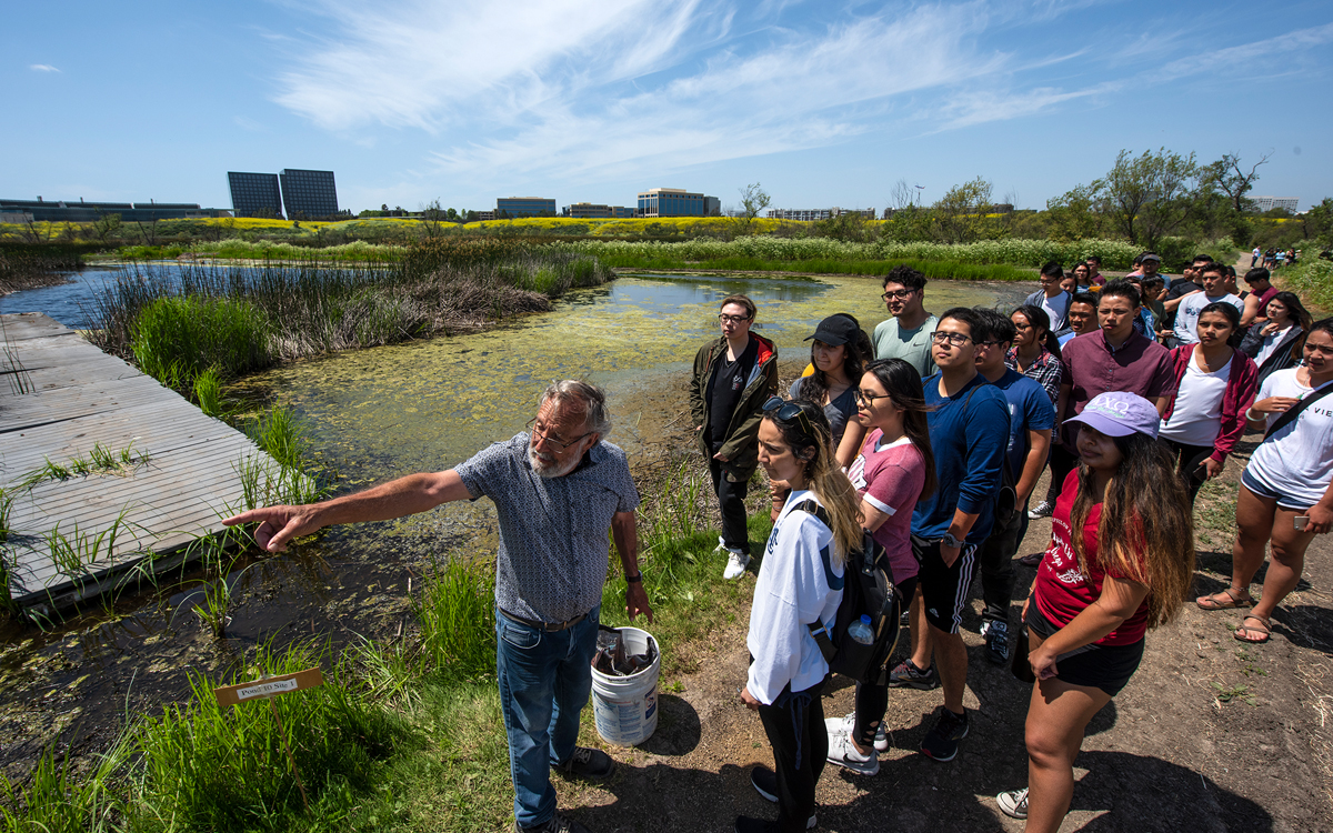 Peter Bowler, standing by a marsh, points to something in the distance while a group of students look on.