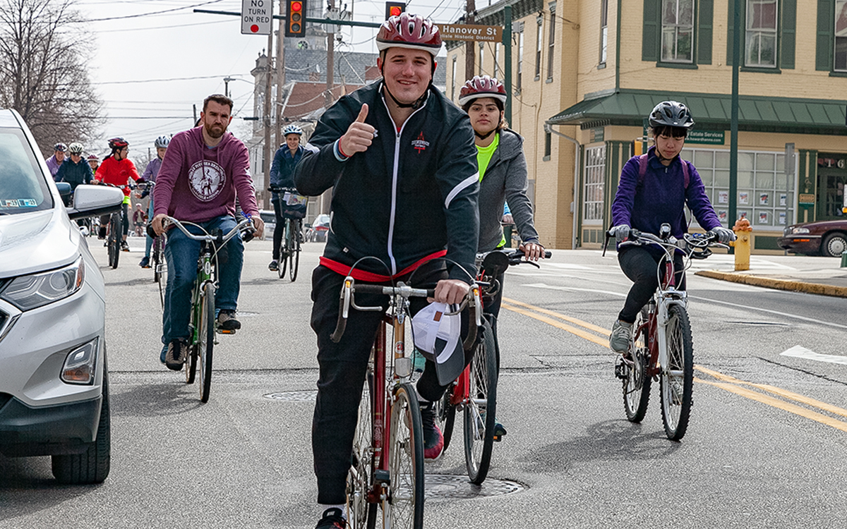 Bicyclists take over a lane, riding toward the camera. A man in front gives a thumbs up and smiles.