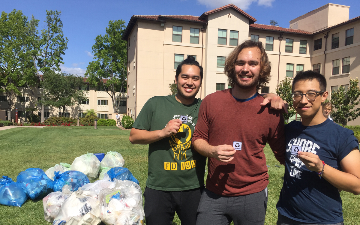 Three male students stand on a lawn next to bags of waste and hold up small white badges with a recycling logo.