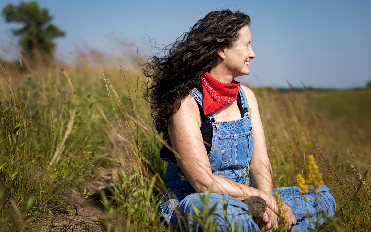 Kelly Madigan is wearing denim overalls and a red handkerchief around her neck. She's sitting in a field and looking off into the distance.
